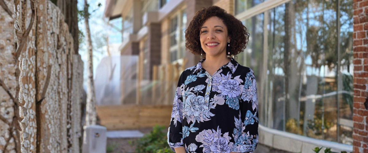 A woman smiles while standing outdoors near a modern building with large windows, trees, and greenery