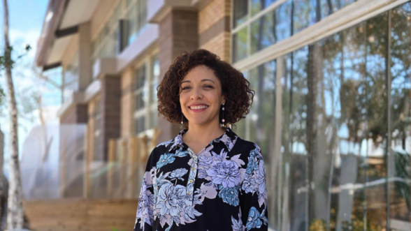 A woman smiles while standing outdoors near a modern building with large windows, trees, and greenery