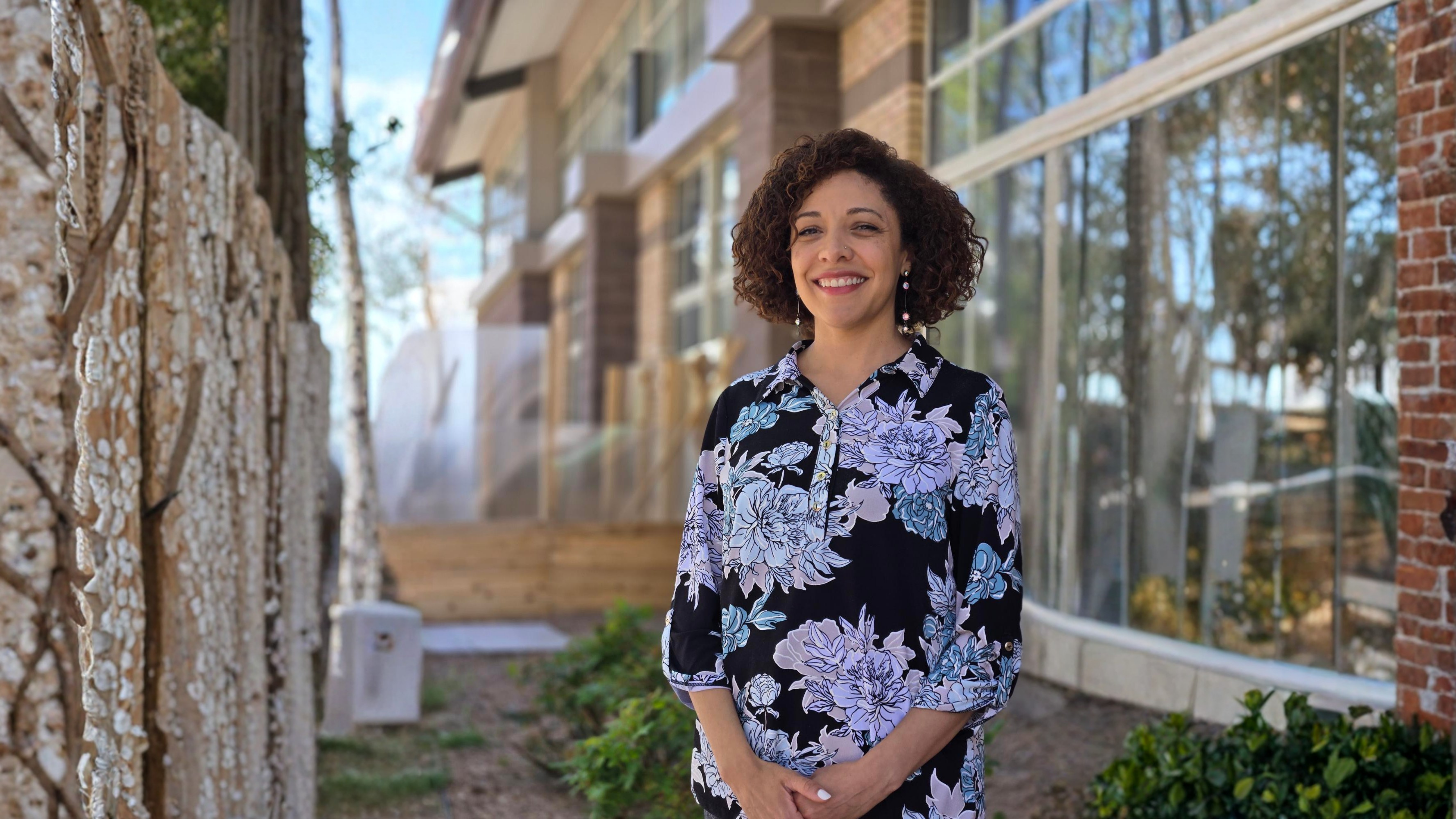 A woman smiles while standing outdoors near a modern building with large windows, trees, and greenery