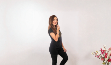 A woman with long brown hair smiles while applying lip gloss, standing against a plain light background and wearing a black T-shirt with gold lettering.