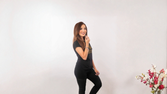 A woman with long brown hair smiles while applying lip gloss, standing against a plain light background and wearing a black T-shirt with gold lettering.