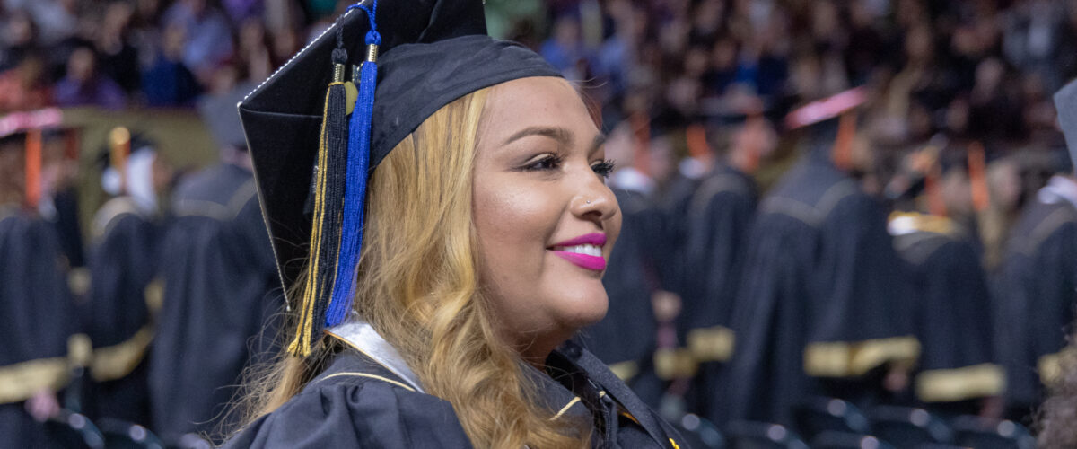 Graduate wearing a cap and gown smiles during a university commencement ceremony, with rows of fellow graduates and a crowded arena in the background.
