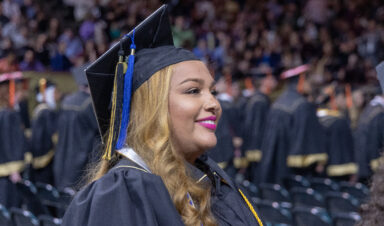 Graduate wearing a cap and gown smiles during a university commencement ceremony, with rows of fellow graduates and a crowded arena in the background.