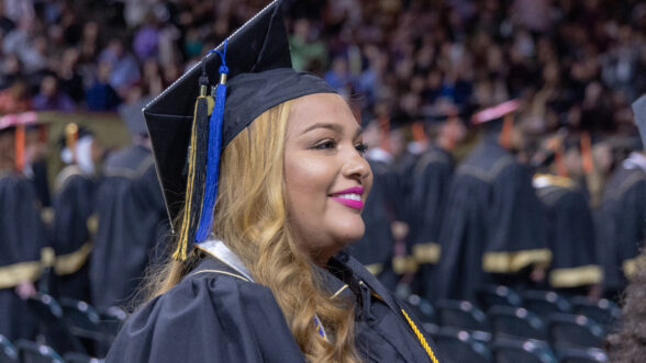 Graduate wearing a cap and gown smiles during a university commencement ceremony, with rows of fellow graduates and a crowded arena in the background.