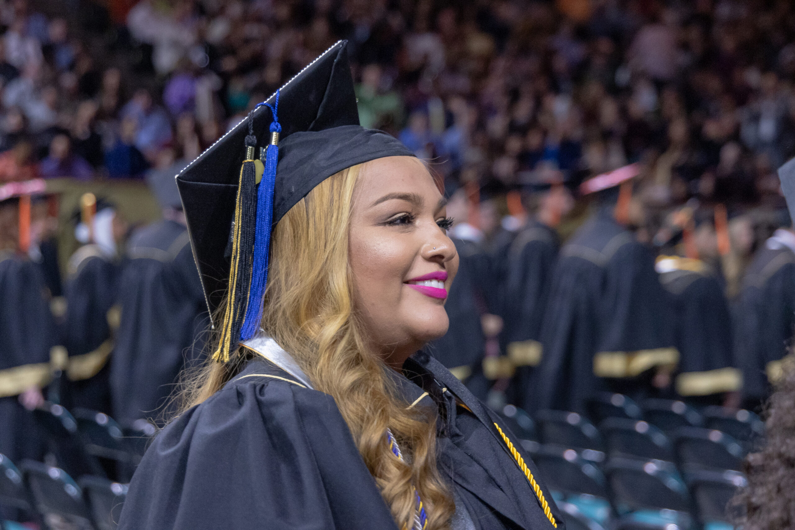 Graduate wearing a cap and gown smiles during a university commencement ceremony, with rows of fellow graduates and a crowded arena in the background.